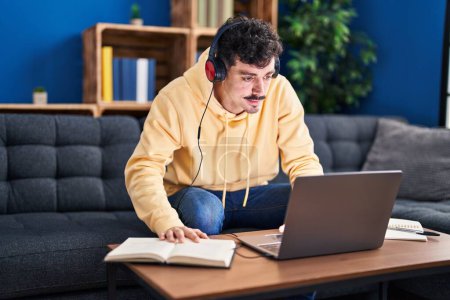 Téléchargez les photos : Jeune homme caucasien utilisant ordinateur portable et écouteurs étudiant à la maison - en image libre de droit