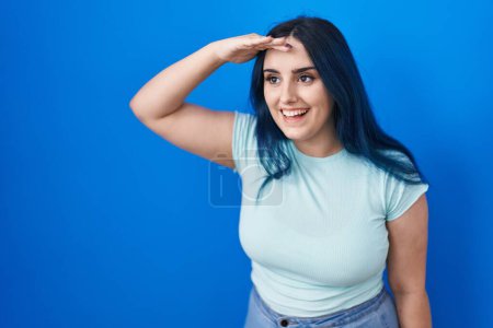 Photo for Young modern girl with blue hair standing over blue background very happy and smiling looking far away with hand over head. searching concept. - Royalty Free Image