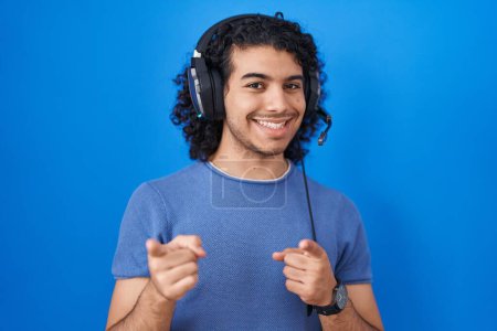 Foto de Hombre hispano con el pelo rizado escuchando música usando auriculares apuntando con los dedos a la cámara con cara alegre y divertida. buena energía y vibraciones. - Imagen libre de derechos
