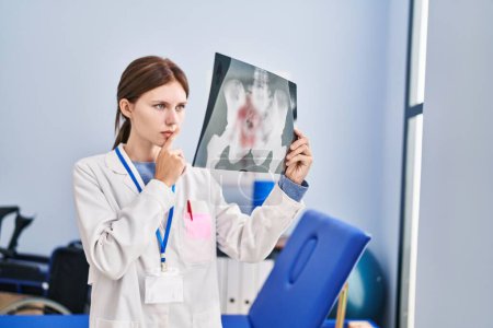 Foto de Joven mujer rubia fisioterapeuta buscando rayos X pensando en la clínica de rehabilitación - Imagen libre de derechos