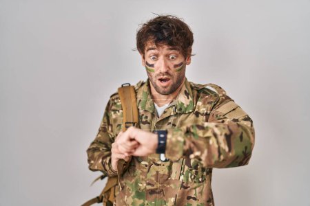 Téléchargez les photos : Jeune homme hispanique portant un uniforme de camouflage de l'armée regardant l'heure de la montre inquiet, peur de se retrouver en retard - en image libre de droit