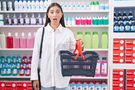 Foto de Mujer hispana joven comprando en farmacia sosteniendo canasta asustada y asombrada con la boca abierta para sorpresa, cara de incredulidad - Imagen libre de derechos