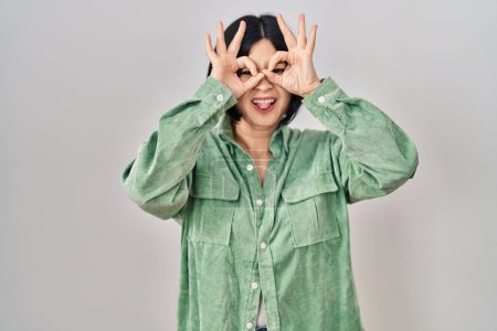 Téléchargez les photos : Jeune femme asiatique debout sur fond blanc faisant un geste correct comme des jumelles sortant la langue, les yeux regardant à travers les doigts. expression folle. - en image libre de droit