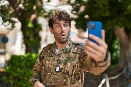 Photo for Hispanic young man wearing camouflage army uniform doing video call scared and amazed with open mouth for surprise, disbelief face - Royalty Free Image