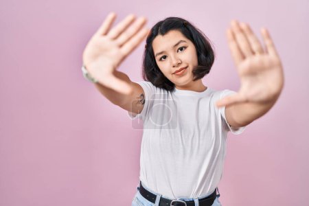 Photo for Young hispanic woman wearing casual white t shirt over pink background doing frame using hands palms and fingers, camera perspective - Royalty Free Image