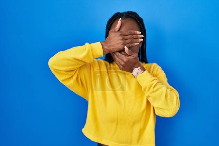 Photo for Beautiful black woman standing over blue background covering eyes and mouth with hands, surprised and shocked. hiding emotion - Royalty Free Image