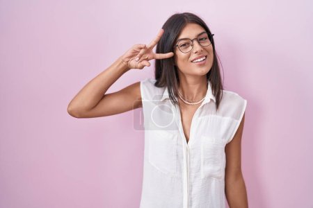 Téléchargez les photos : Brunette jeune femme debout sur fond rose portant des lunettes faisant symbole de paix avec les doigts sur le visage, souriant joyeuse montrant la victoire - en image libre de droit