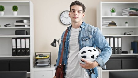 Foto de Joven trabajador de negocios hispano sosteniendo casco de bicicleta con expresión relajada en la oficina - Imagen libre de derechos