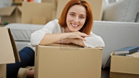 Photo for Young redhead woman leaning on cardboard box holding keys smiling at new home - Royalty Free Image