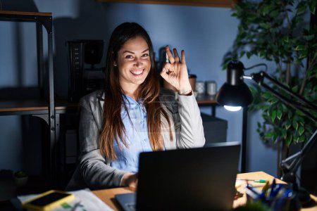 Photo for Young brunette woman working at the office at night smiling positive doing ok sign with hand and fingers. successful expression. - Royalty Free Image