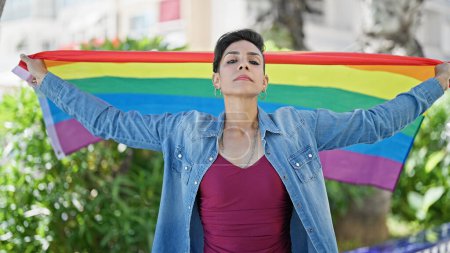 Foto de Joven mujer hispana hermosa de pie con expresión seria sosteniendo la bandera del arco iris en el parque - Imagen libre de derechos