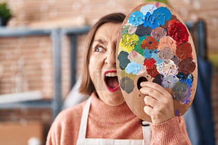 Photo for Middle age hispanic woman holding painter palette close to face angry and mad screaming frustrated and furious, shouting with anger. rage and aggressive concept. - Royalty Free Image