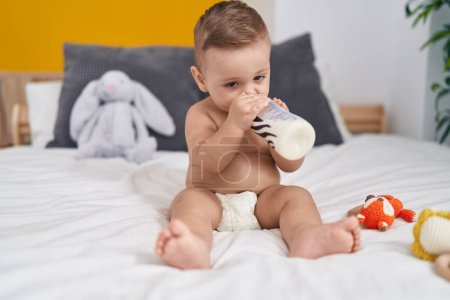 Téléchargez les photos : Adorable bébé caucasien suçant le lait sur le biberon assis sur le lit dans la chambre - en image libre de droit