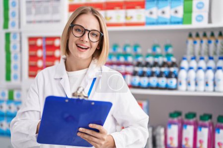 Téléchargez les photos : Jeune femme blonde pharmacienne souriante écriture confiante sur le document à la pharmacie - en image libre de droit