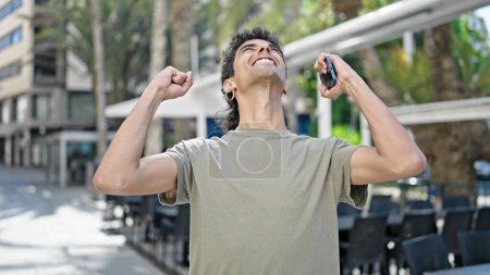 Téléchargez les photos : Jeune homme hispanique utilisant un smartphone avec geste gagnant sur la terrasse du café - en image libre de droit
