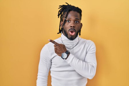 Photo for African man with dreadlocks wearing turtleneck sweater over yellow background surprised pointing with finger to the side, open mouth amazed expression. - Royalty Free Image