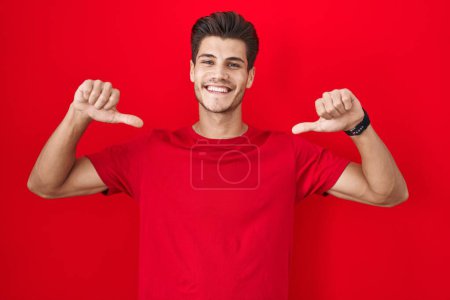 Foto de Joven hombre hispano de pie sobre fondo rojo mirando confiado con sonrisa en la cara, señalándose con los dedos orgullosos y felices. - Imagen libre de derechos