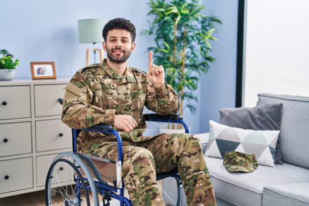 Téléchargez les photos : Arabe homme portant uniforme de camouflage de l'armée assis sur fauteuil roulant montrant et pointant vers le haut avec le doigt numéro un tout en souriant confiant et heureux. - en image libre de droit