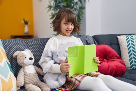 Photo for Adorable hispanic girl reading book sitting on sofa at home - Royalty Free Image