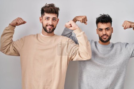 Téléchargez les photos : Jeune couple homosexuel debout sur fond blanc montrant les muscles des bras souriant fier. concept de fitness. - en image libre de droit