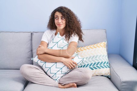 Foto de Mujer hispana con el pelo rizado sentada en el sofá en casa sonriendo mirando a un lado y mirando hacia otro pensando. - Imagen libre de derechos
