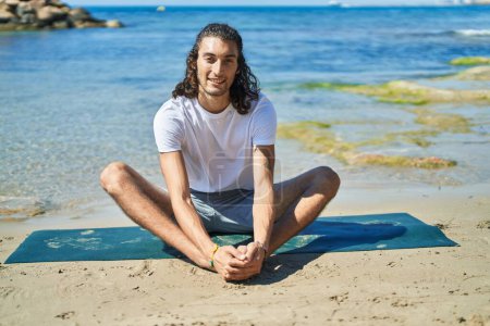 Téléchargez les photos : Jeune homme hispanique faisant de l'exercice de yoga assis sur le sable à la plage - en image libre de droit