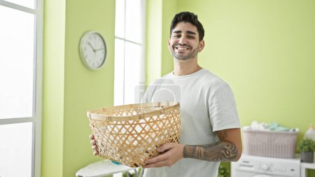 Foto de Joven hombre hispano sonriendo confiado sosteniendo cesta con ropa en la lavandería - Imagen libre de derechos