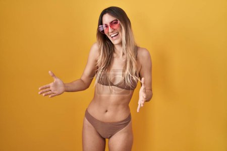Foto de Mujer hispana joven vistiendo bikini sobre fondo amarillo sonriente alegre con los brazos abiertos como bienvenida amistosa, saludos positivos y confiados - Imagen libre de derechos