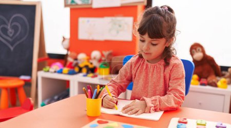 Foto de Adorable estudiante hispana sentada en la mesa dibujando en un cuaderno en el jardín de infantes - Imagen libre de derechos