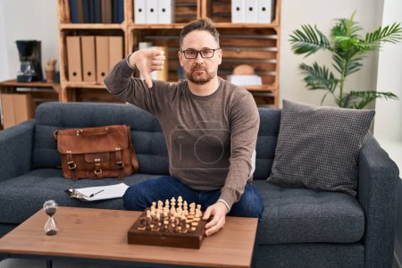 Photo for Middle age caucasian man playing chess sitting on the sofa with angry face, negative sign showing dislike with thumbs down, rejection concept - Royalty Free Image