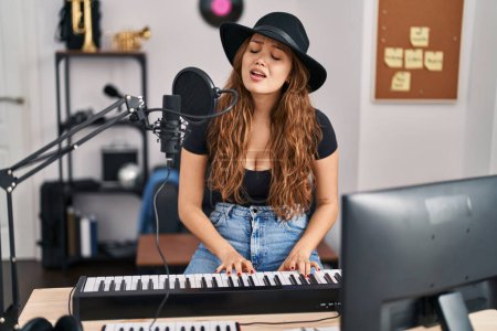 Foto de Joven hermosa mujer hispana músico cantando canción tocando el piano en el estudio de música - Imagen libre de derechos