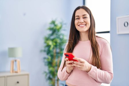 Téléchargez les photos : Jeune femme souriant confiant en utilisant un smartphone à la maison - en image libre de droit