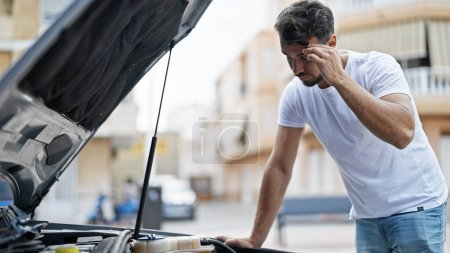 Téléchargez les photos : Jeune homme hispanique regardant voiture à moteur avec expression inquiète à la rue - en image libre de droit
