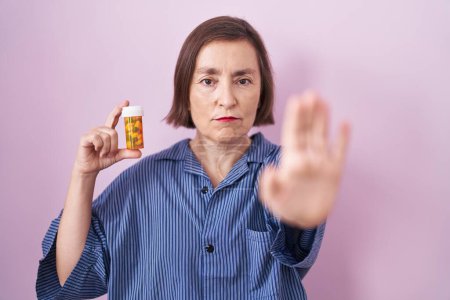Foto de Middle age hispanic woman holding pills with open hand doing stop sign with serious and confident expression, defense gesture - Imagen libre de derechos