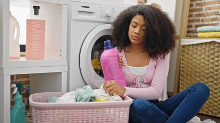Téléchargez les photos : Femme afro-américaine laver les vêtements tenant bouteille de détergent avec expression triste à la buanderie - en image libre de droit