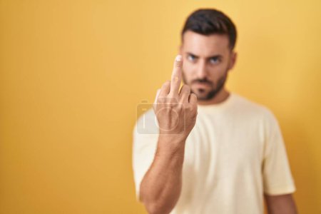 Photo for Handsome hispanic man standing over yellow background showing middle finger, impolite and rude fuck off expression - Royalty Free Image