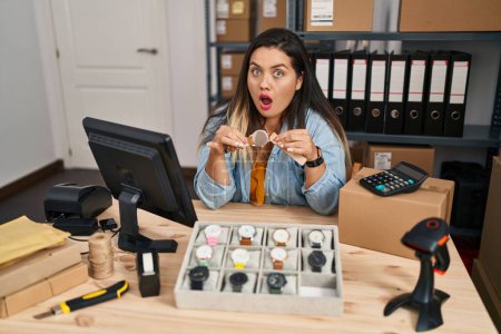 Foto de Mujer hispana joven que trabaja en el comercio electrónico de pequeñas empresas vendiendo relojes asustada y sorprendida con la expresión sorpresa y asombrada, el miedo y la cara emocionada. - Imagen libre de derechos