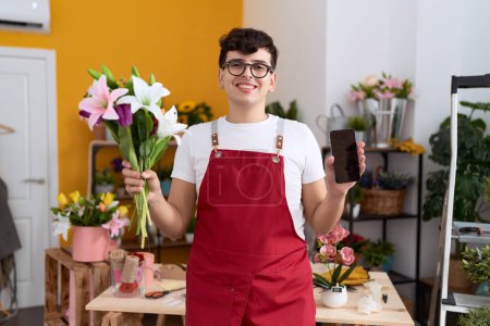 Téléchargez les photos : Jeune homme non binaire travaillant à la boutique de fleuristes montrant écran smartphone souriant avec un sourire heureux et cool sur le visage. montrant les dents. - en image libre de droit