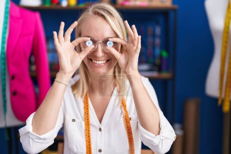 Téléchargez les photos : Young blonde woman tailor smiling confident holding buttons over eyes at sewing studio - en image libre de droit