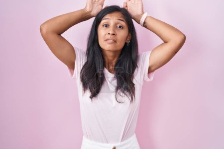 Foto de Young hispanic woman standing over pink background doing bunny ears gesture with hands palms looking cynical and skeptical. easter rabbit concept. - Imagen libre de derechos
