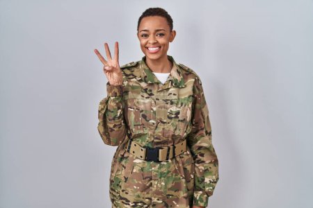 Foto de Hermosa mujer afroamericana vistiendo camuflaje uniforme del ejército mostrando y señalando con los dedos número tres mientras sonríe confiado y feliz. - Imagen libre de derechos