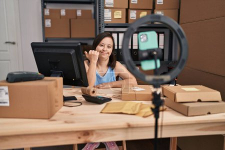 Photo for Hispanic girl with down syndrome working at small business ecommerce doing online tutorial pointing thumb up to the side smiling happy with open mouth - Royalty Free Image