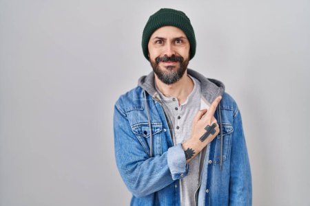 Téléchargez les photos : Jeune homme hispanique avec des tatouages portant un bonnet de laine gai avec un sourire du visage pointant avec la main et le doigt sur le côté avec une expression heureuse et naturelle sur le visage - en image libre de droit