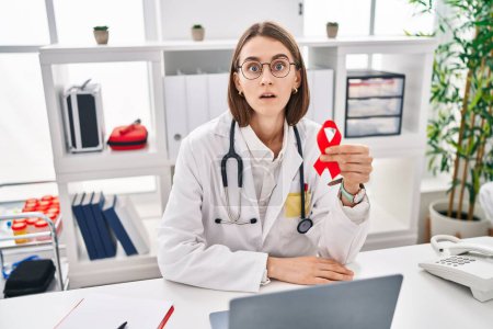 Photo for Young caucasian doctor woman holding support red ribbon scared and amazed with open mouth for surprise, disbelief face - Royalty Free Image