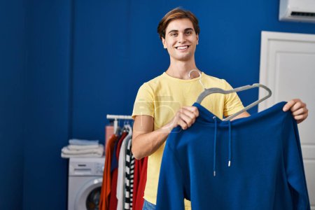 Photo for Young caucasian man smiling confident putting sweatshirt on hanger at laundry room - Royalty Free Image