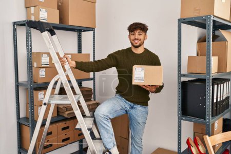 Photo for Young arab man ecommerce business worker holding package standing on ladder at office - Royalty Free Image