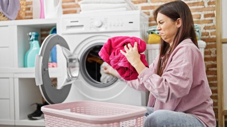 Téléchargez les photos : Jeune belle femme hispanique ouvrant la machine à laver sentant les vêtements sales à la buanderie - en image libre de droit