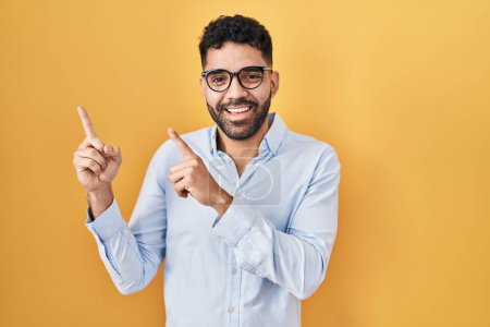 Téléchargez les photos : Hispanic man with beard standing over yellow background smiling and looking at the camera pointing with two hands and fingers to the side. - en image libre de droit
