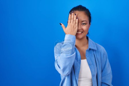 Téléchargez les photos : Asiatique jeune femme debout sur fond bleu couvrant un oeil avec la main, sourire confiant sur le visage et l'émotion surprise. - en image libre de droit