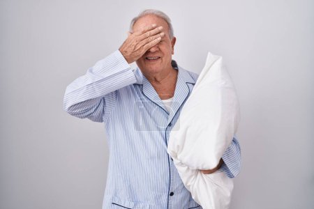 Photo for Senior man with grey hair wearing pijama hugging pillow smiling and laughing with hand on face covering eyes for surprise. blind concept. - Royalty Free Image
